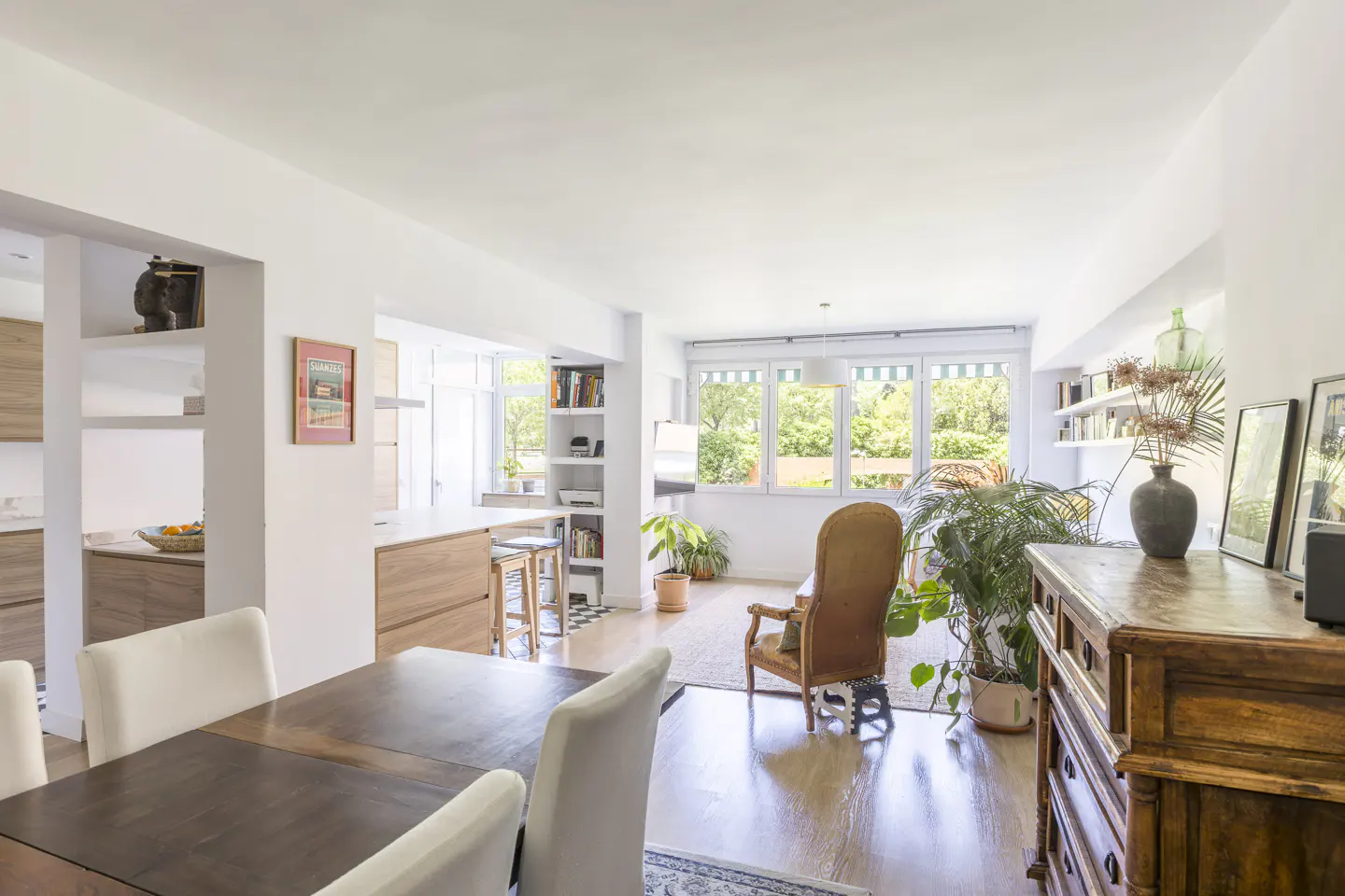 Bright, open-plan living space with a dining table, chairs, kitchen, and a cozy armchair near large windows. Plants add a touch of nature.