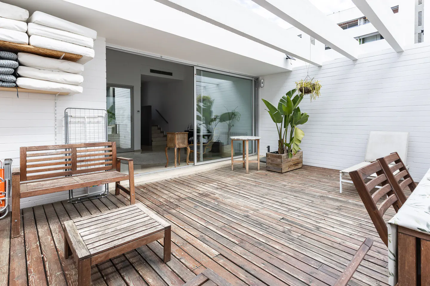 Outdoor patio with wooden deck, white walls, and sliding glass doors. Furniture includes a wooden bench, table, chairs, and potted plants.