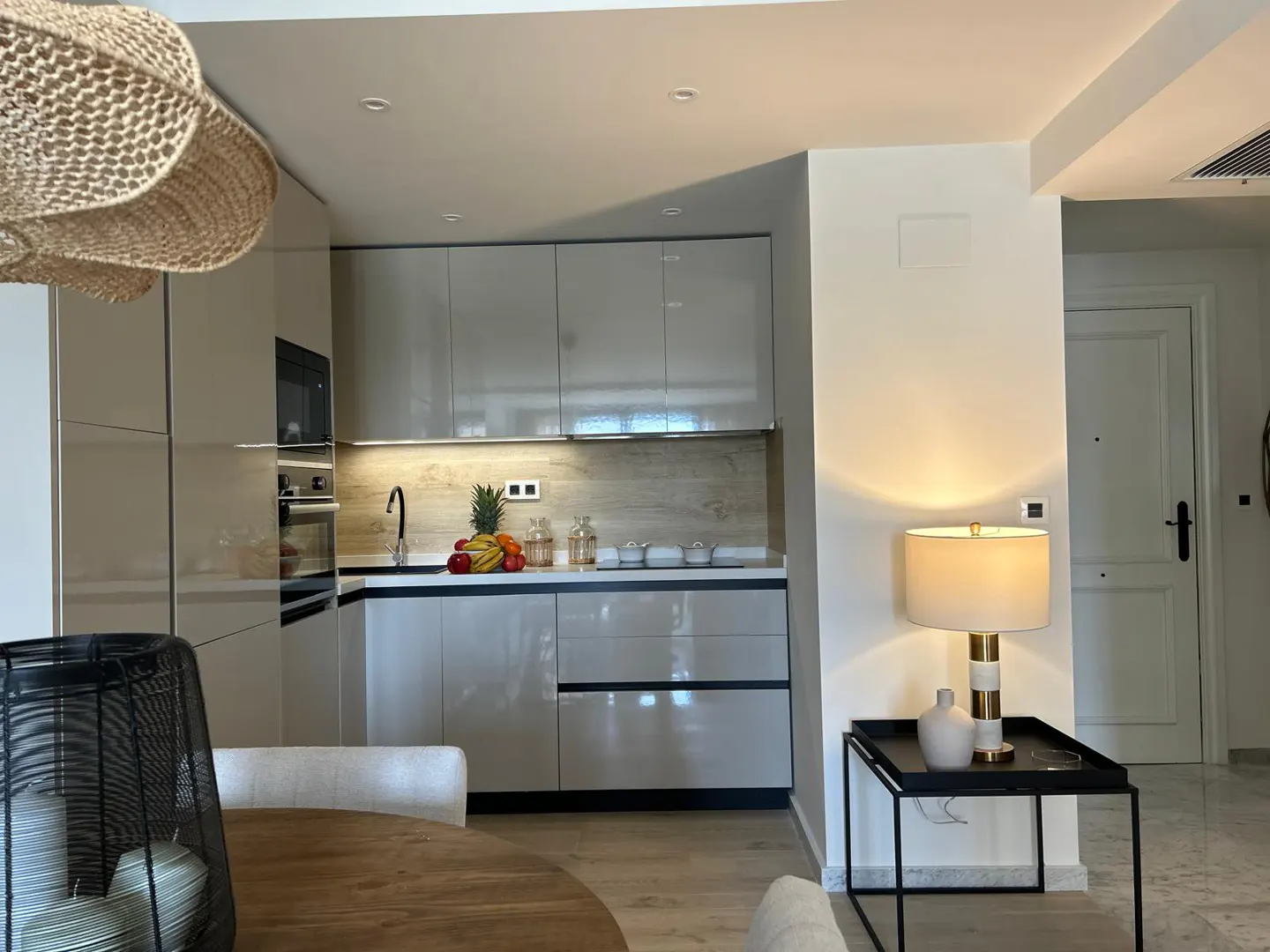 A modern kitchen with glossy cabinets, a black faucet, and fruit on the counter. A lamp sits on a black table near a white door.