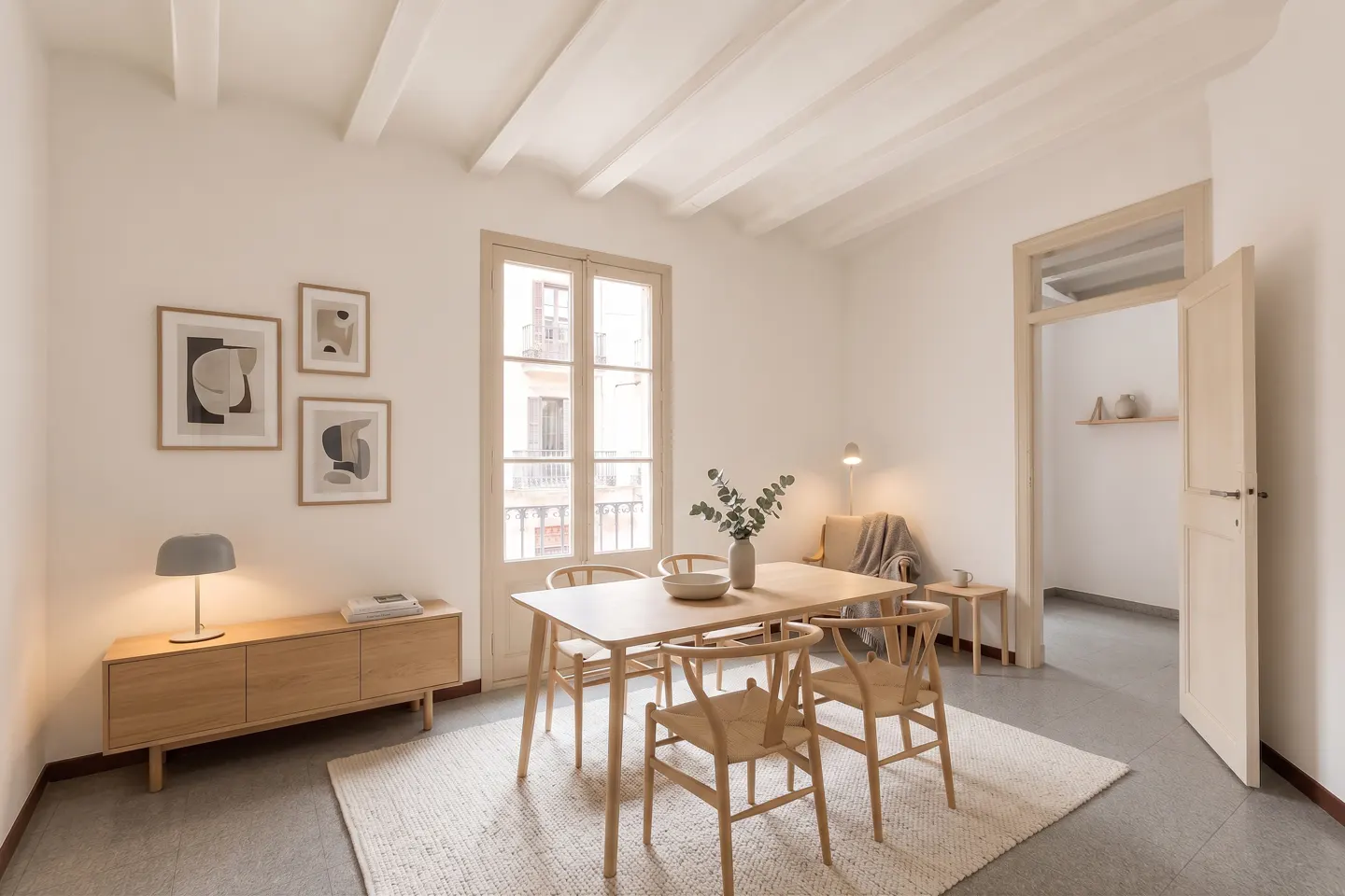 Bright dining room with a light wood table, four chairs, and a sideboard on a white rug. Art on the wall and a window with a balcony.