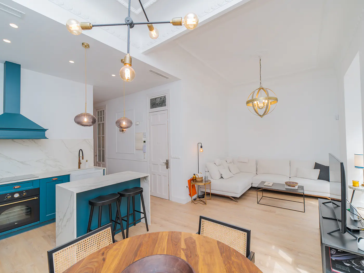 Bright, open-concept living space with a blue kitchen, island with stools, and a round wooden table with chairs. A white sofa and TV are in the background.