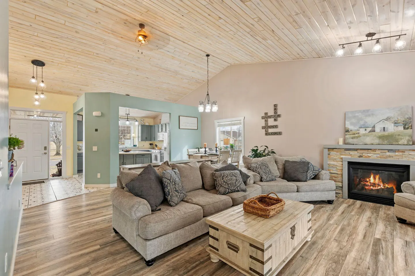 Living room with wood floors, a stone fireplace, and a beige sectional sofa. A wooden chest sits in front of the sofa. The ceiling is wood paneling.