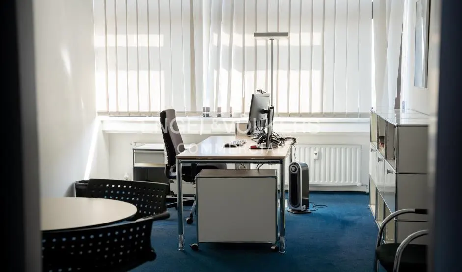 Bright office space with a desk, computer, chair, and blue carpet. White blinds cover the window.