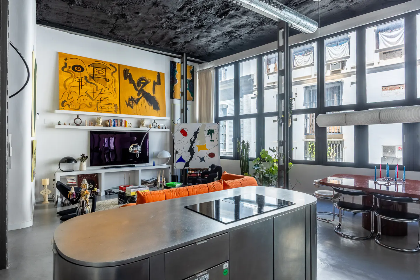 Modern apartment interior with stainless steel kitchen island, orange sofa, and large windows. Abstract art adorns the walls.