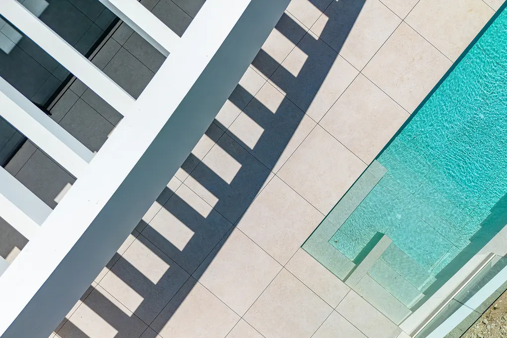 Aerial view of a turquoise pool with steps, beige tile patio, and white pergola casting shadows.