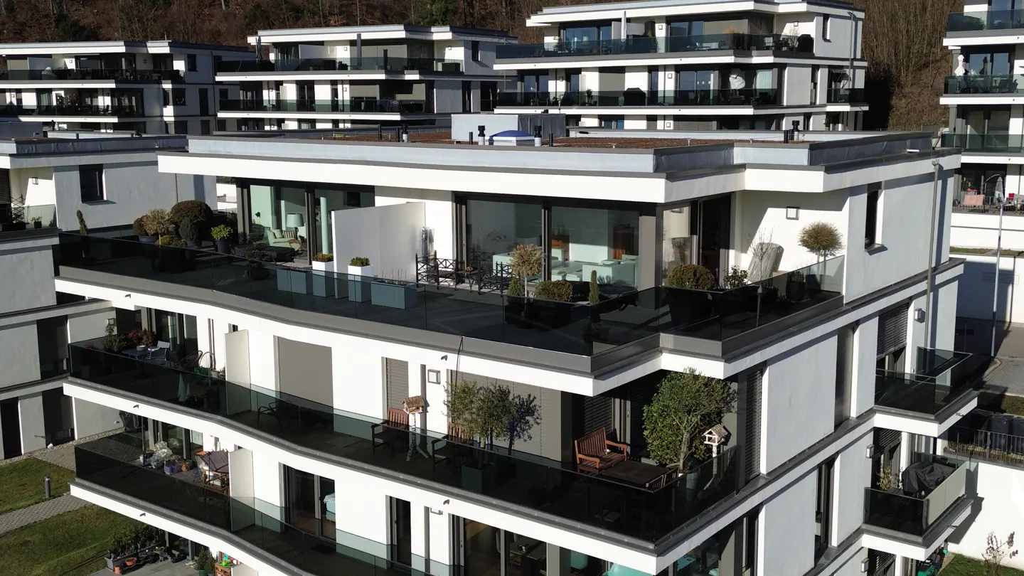 Modern white apartment building with glass balconies and outdoor furniture, set against a backdrop of trees.