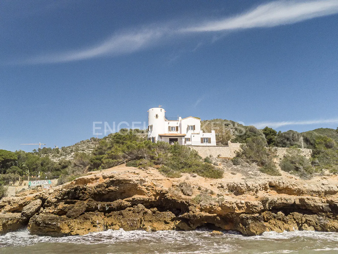White house with a tower on a rocky cliff overlooking the ocean under a blue sky.