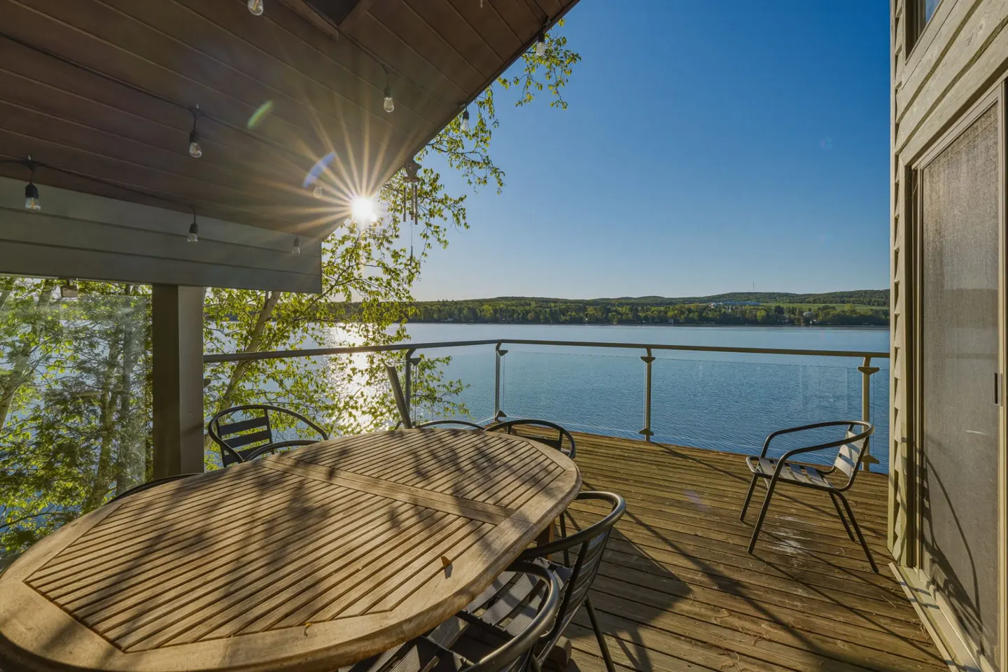 Outdoor wooden deck with table, chairs, and glass railing overlooking a blue lake and green trees under a sunny blue sky.