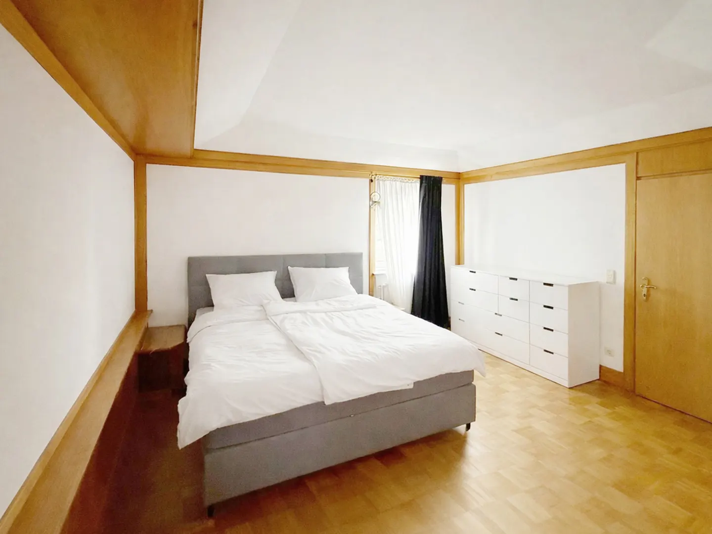 Bright bedroom with a gray bed, white linens, and a white dresser. Wood trim accents the walls and ceiling. Hardwood floors add warmth.