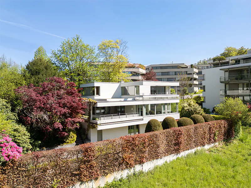 Modern white house with balconies, surrounded by lush greenery and a trimmed hedge under a clear blue sky.