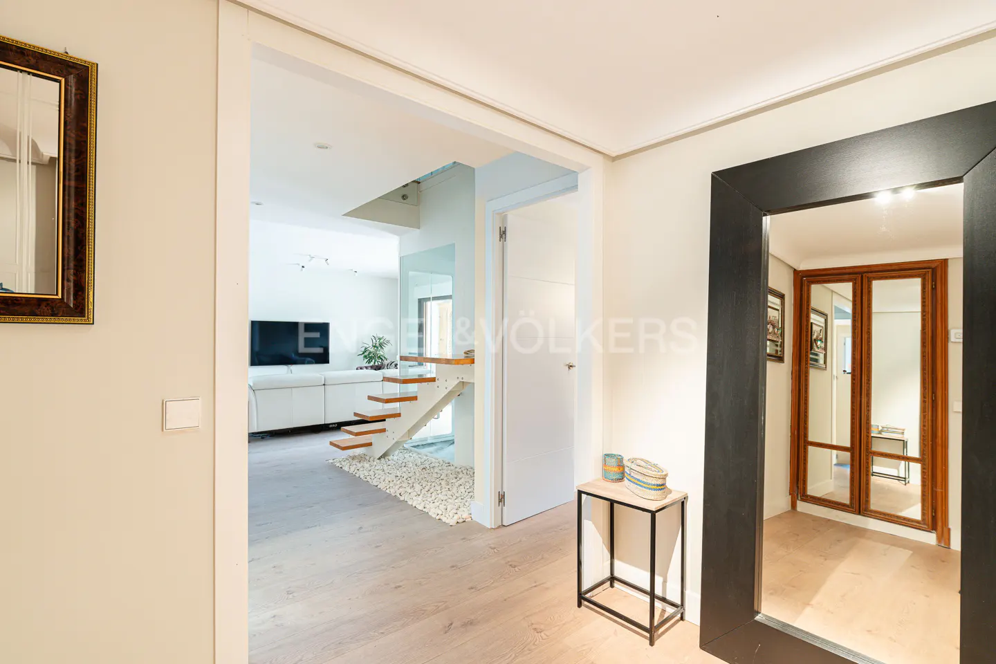Hallway view of a modern home with light wood floors, white walls, and a staircase with glass railings. A black-framed mirror reflects a wood-framed door.