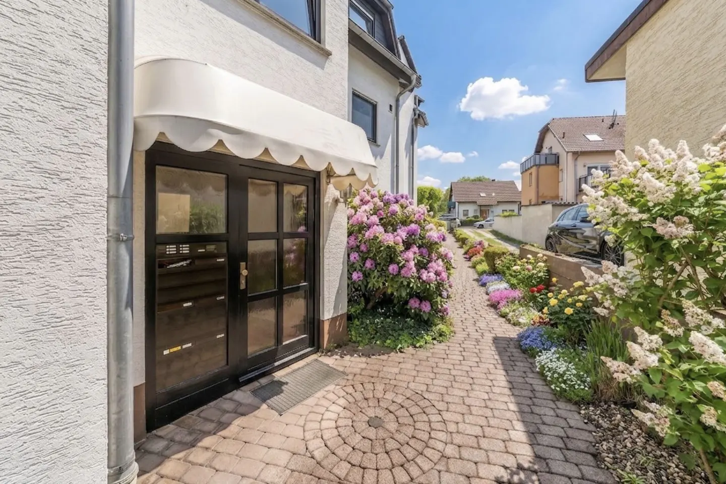 House entrance with black door, white awning, and brick path lined with colorful flowers and greenery.