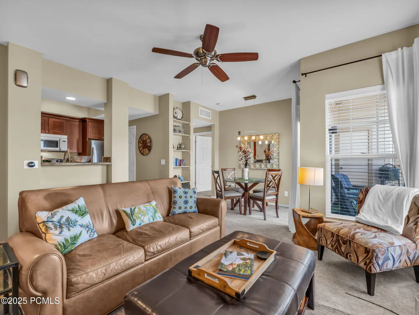 Living room with brown leather sofa, ottoman, patterned chair, and dining table. A ceiling fan hangs above. Kitchen visible in the background.