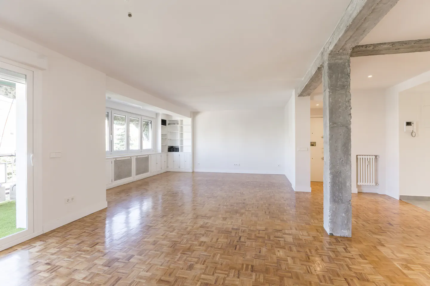 Spacious, empty living room with white walls, parquet wood flooring, and a prominent exposed concrete support pillar.
