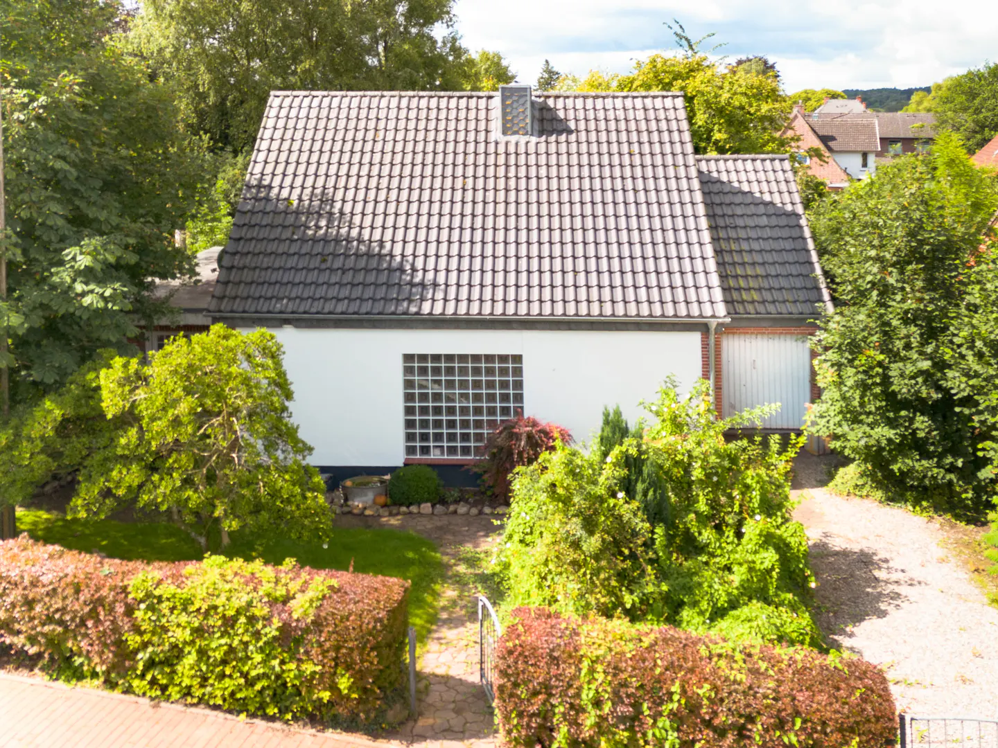 A white house with a gray tiled roof, surrounded by green trees and bushes. A brick path leads to the front door.