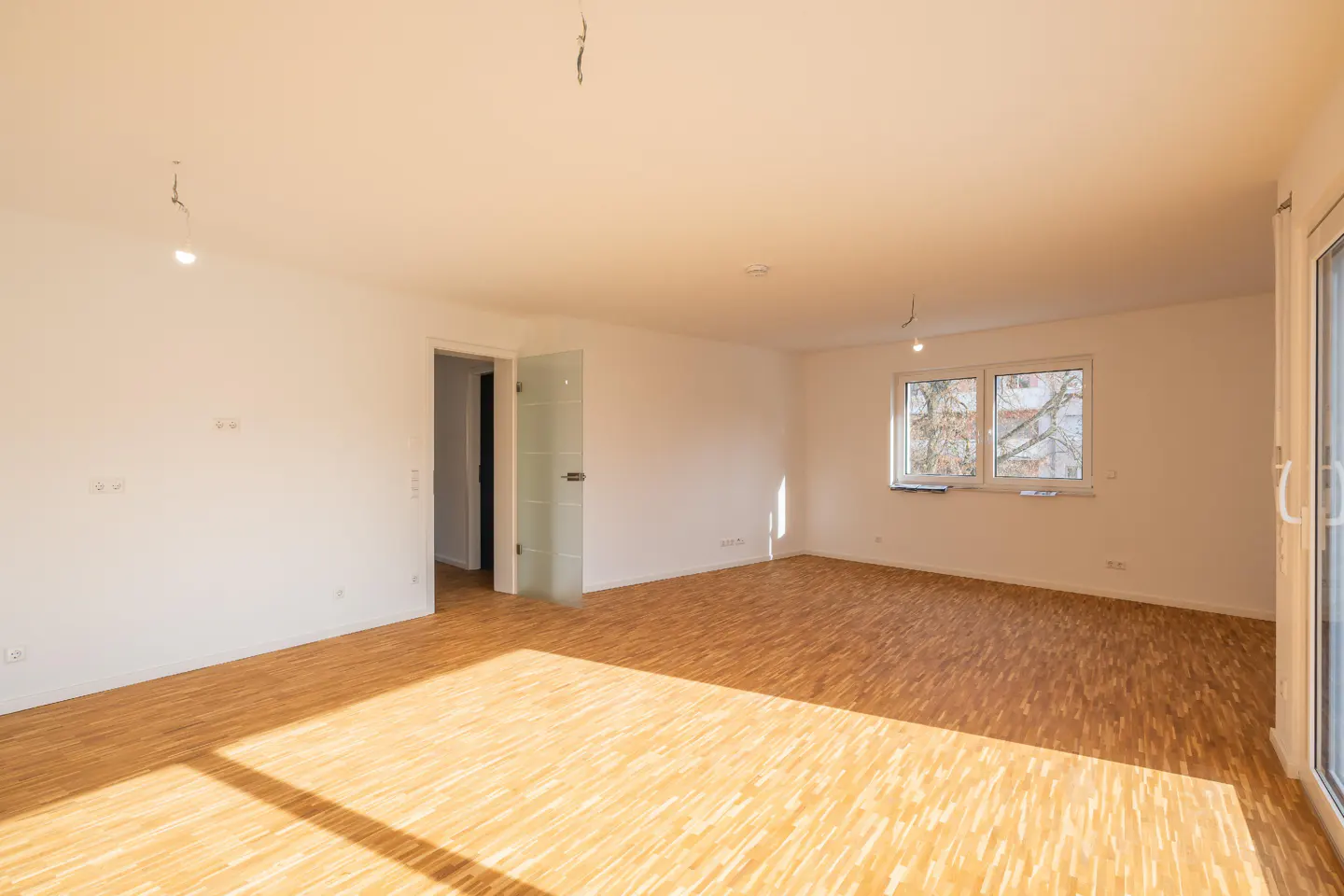 Bright, empty room with light wood floors, white walls, a window, and a glass door. Sunlight streams across the floor.