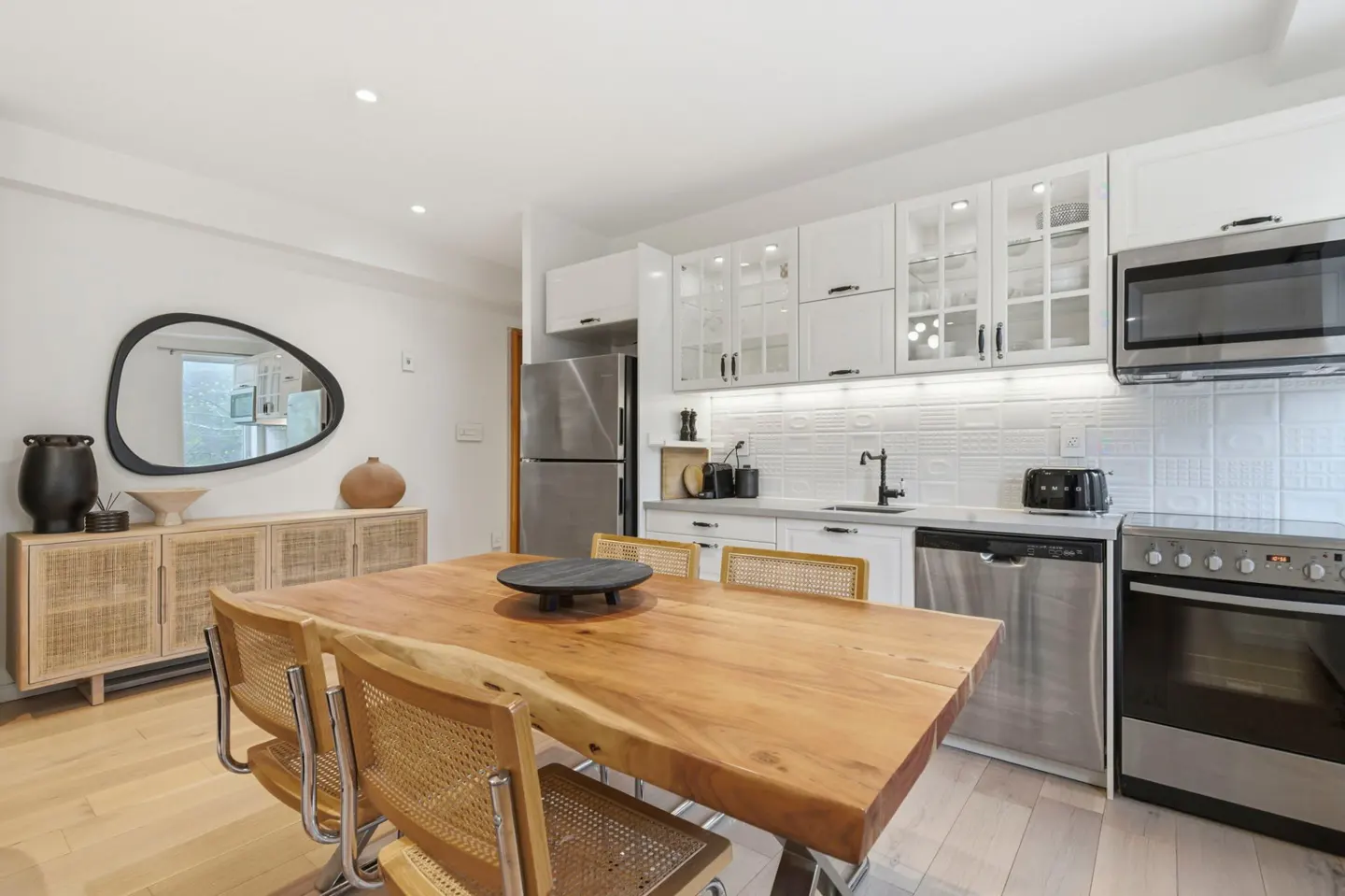 Bright kitchen with a wooden table, wicker chairs, and white cabinets. Stainless steel appliances and a unique mirror add modern touches.