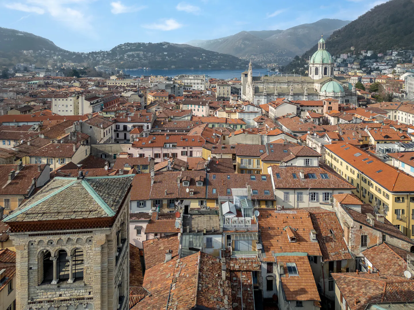 Aerial view of Como, Italy, featuring red-tiled roofs, a cathedral with a green dome, and mountains in the background.