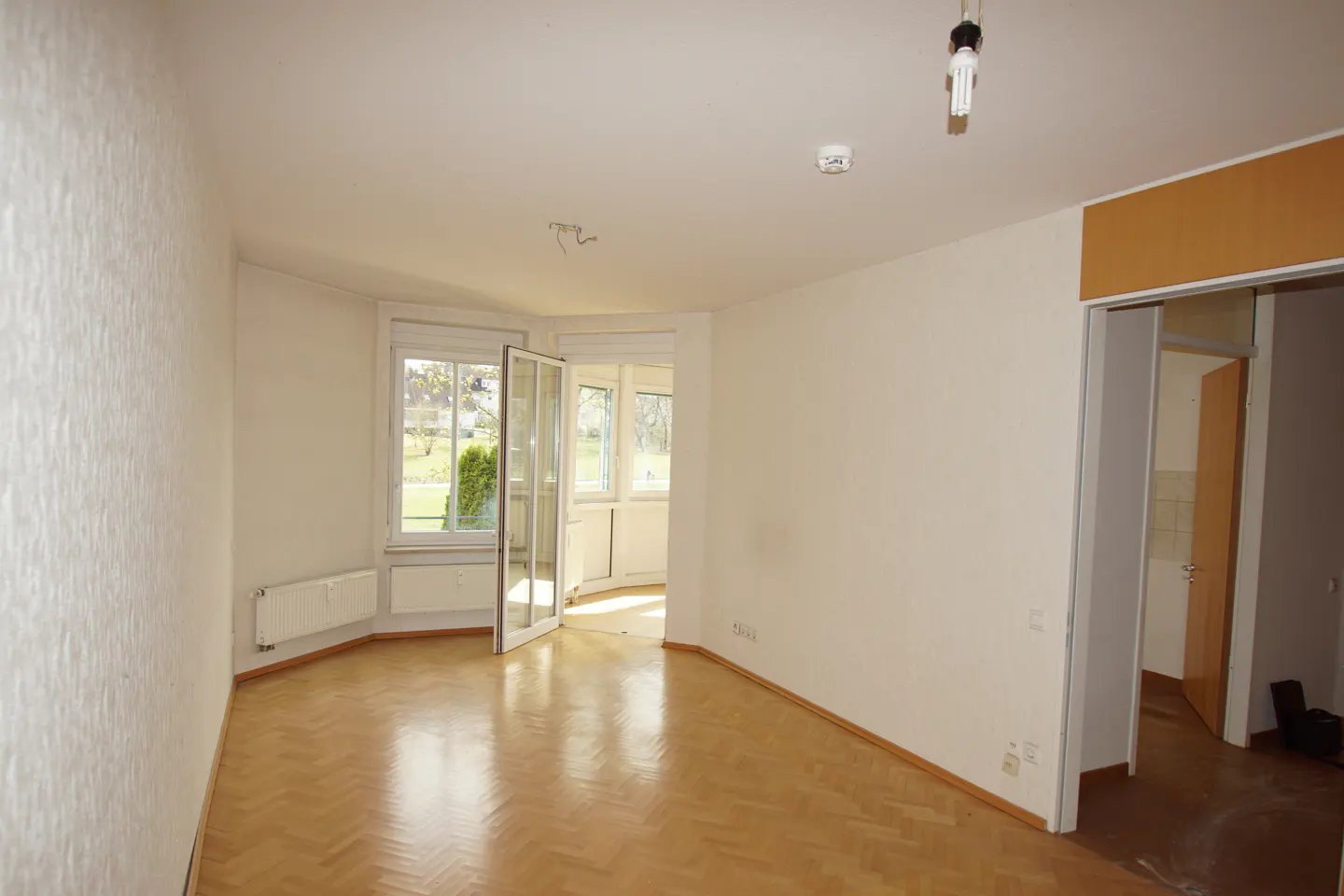 Bright, empty room with parquet flooring, white walls, and a window with a view of greenery. A doorway leads to another room.