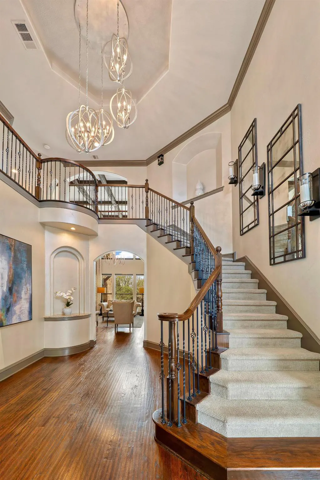 Grand foyer with wood floors, staircase, and wrought iron railings. Three modern chandeliers hang from the ceiling. Two mirrors adorn the wall.