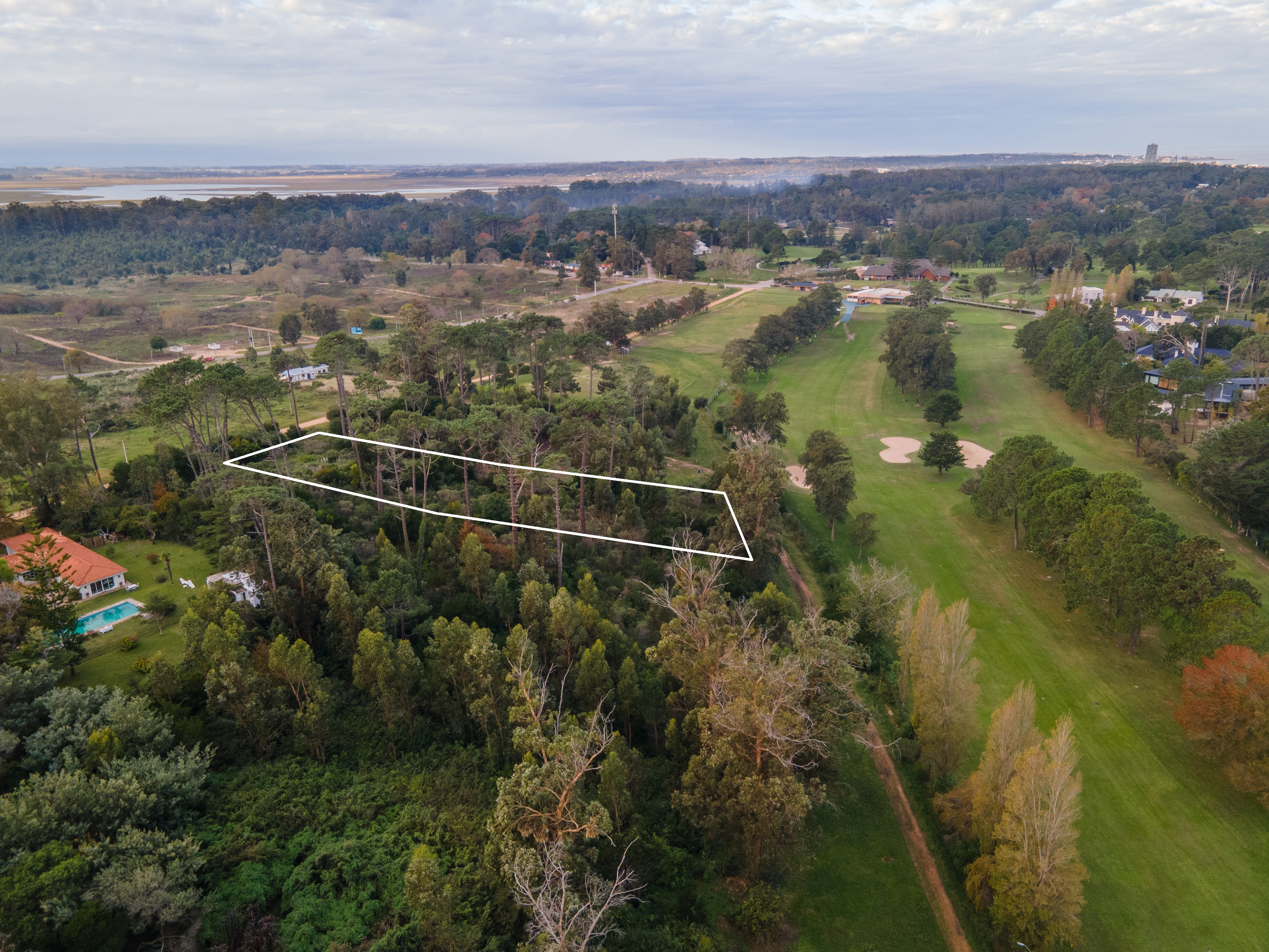 Aerial view of a wooded lot outlined in white, adjacent to a golf course and residential area under a cloudy sky.