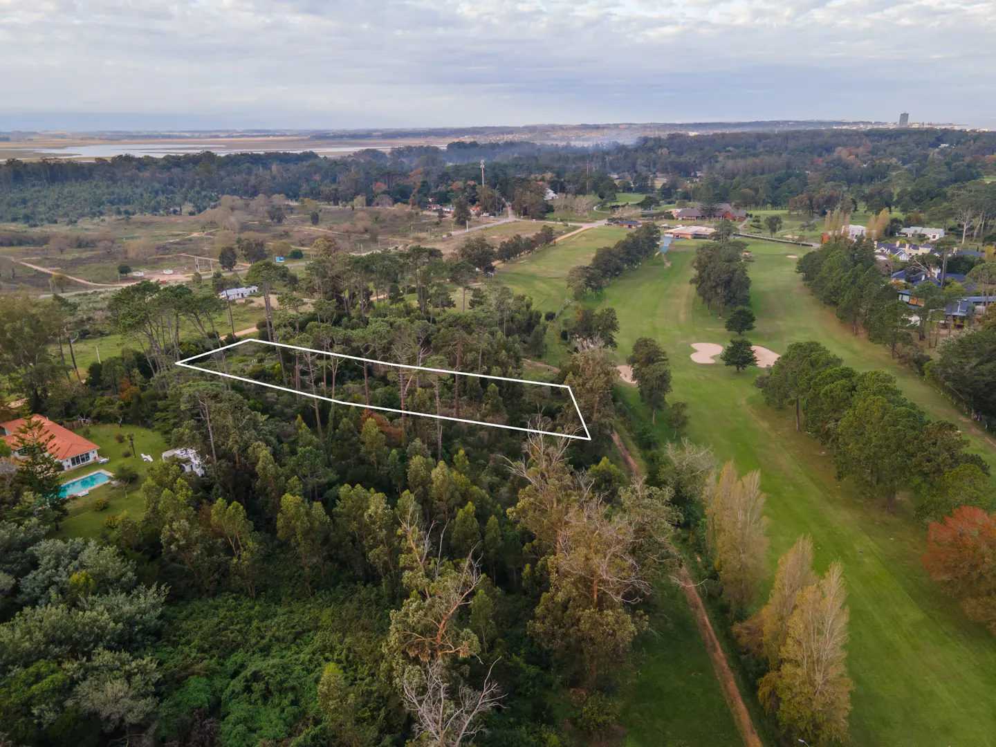 Aerial view of a wooded lot outlined in white, adjacent to a golf course and residential area under a cloudy sky.