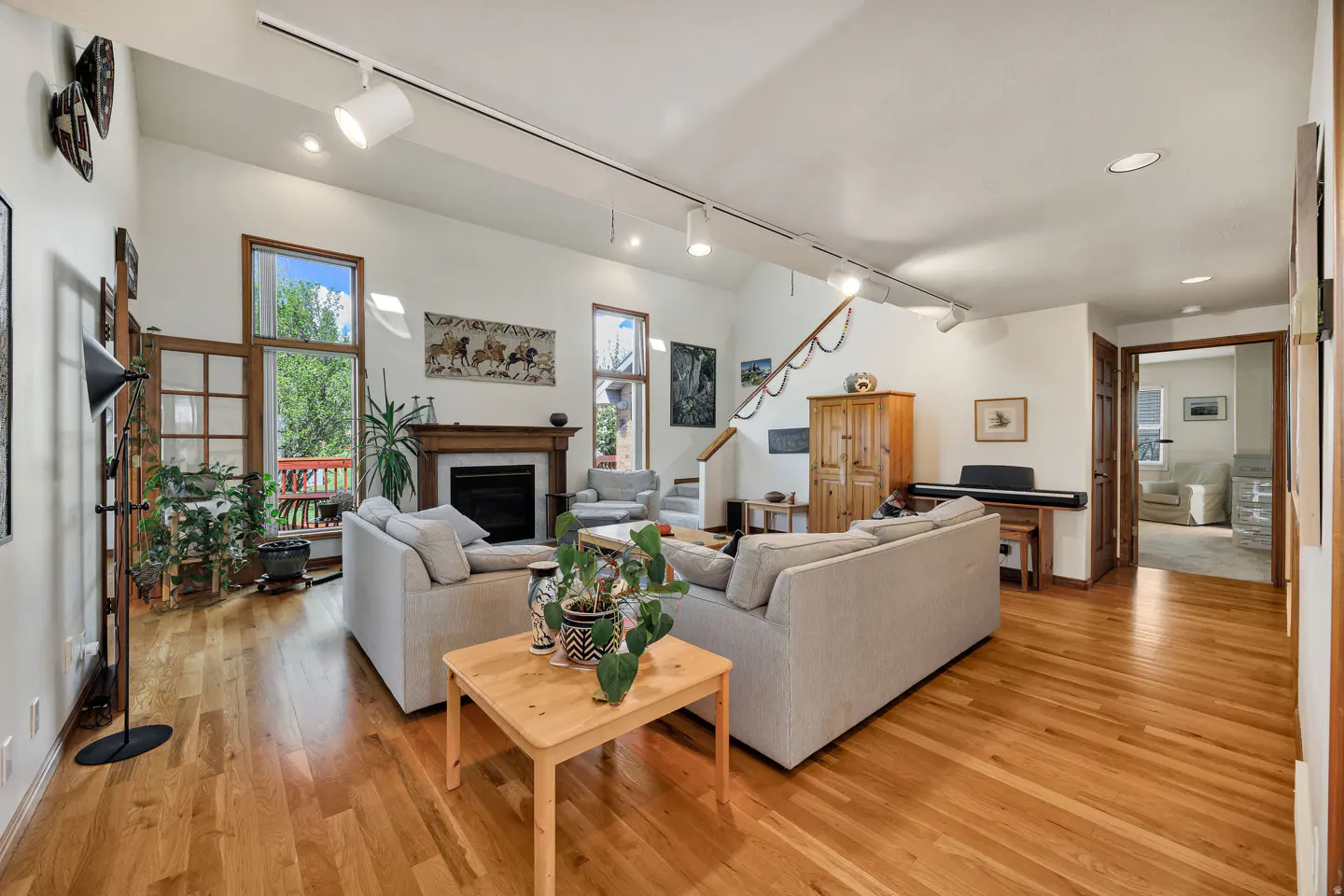 Bright living room with hardwood floors, white walls, and track lighting. Two sofas face each other with a wood table in between. A fireplace, piano, and staircase are visible.