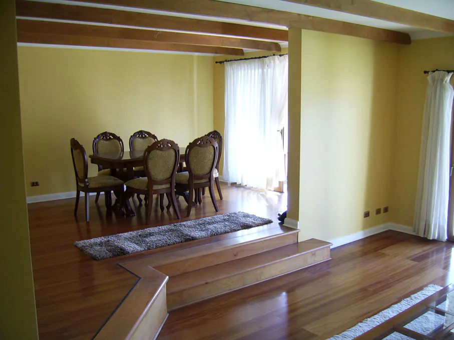 Interior view of a dining room with wooden floors, yellow walls, and a wooden table with six chairs. A rug sits on the steps leading to the dining area. White curtains hang in the background.