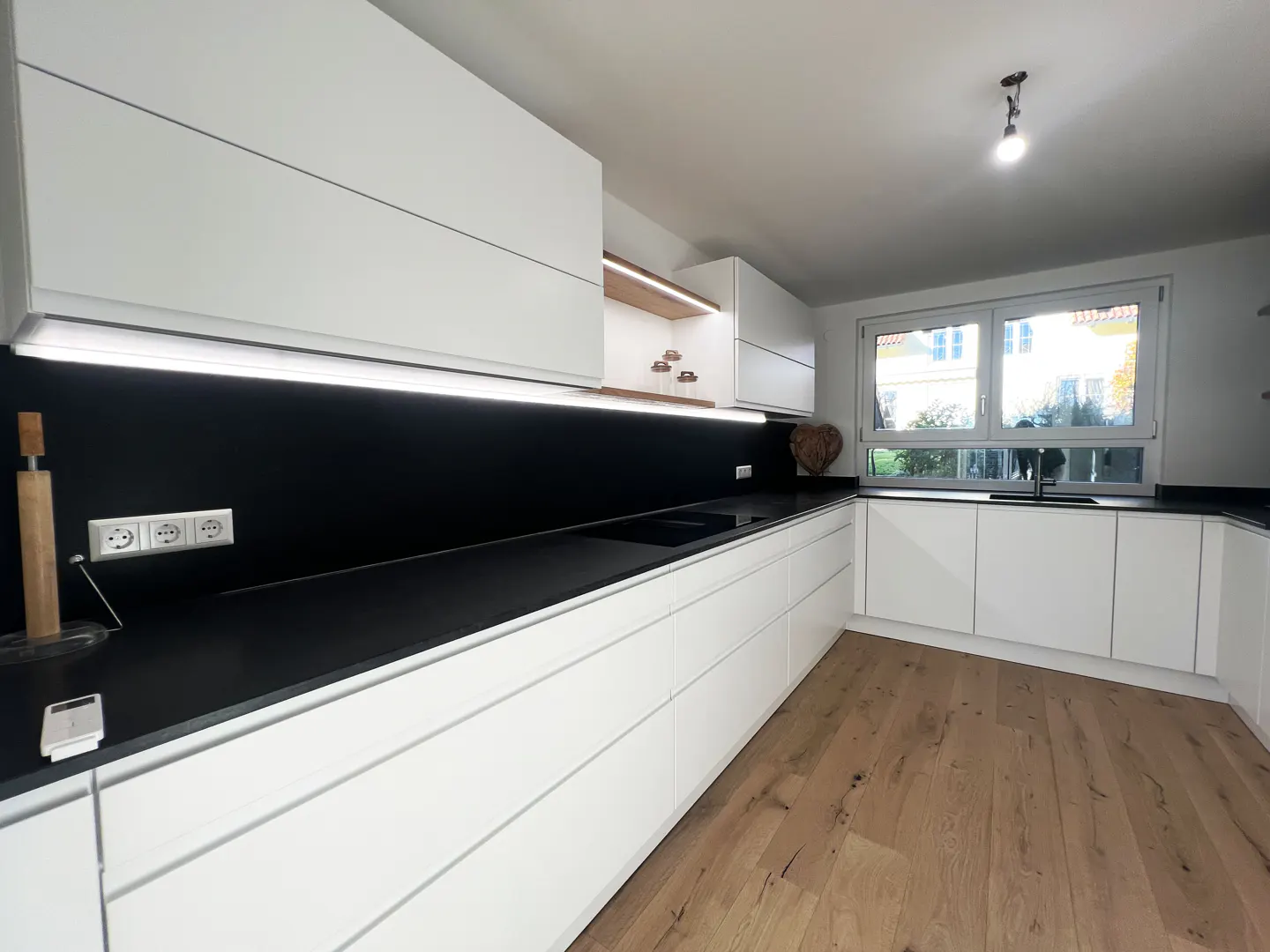 A modern kitchen with white cabinets, black countertops, and wood floors. A window provides natural light.