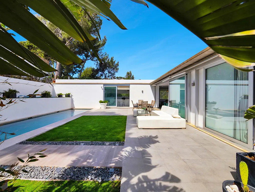 Modern patio with a white sofa, dining set, and swimming pool next to a green lawn under a bright blue sky.