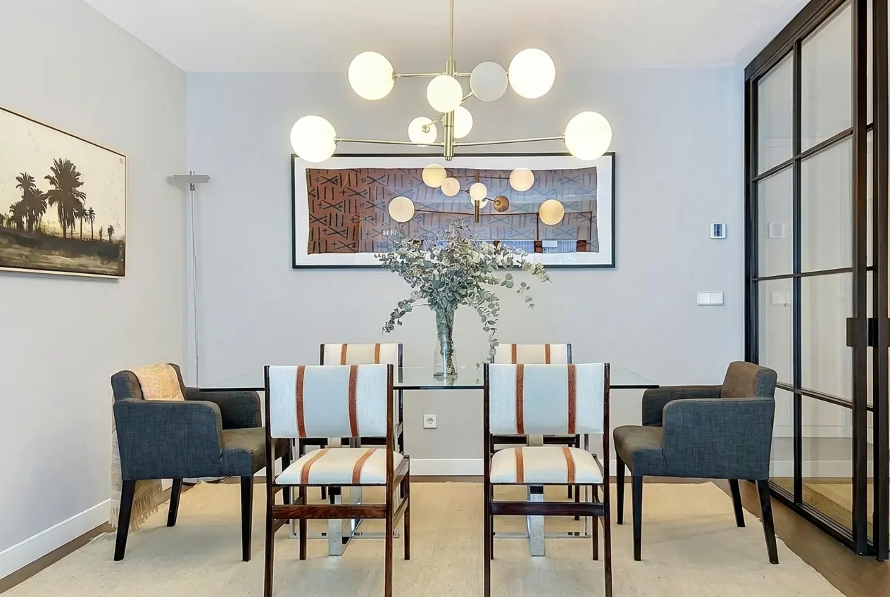 Dining room with a glass table, six chairs, and a modern chandelier. Artwork hangs on the light gray walls. A black framed glass door is on the right.