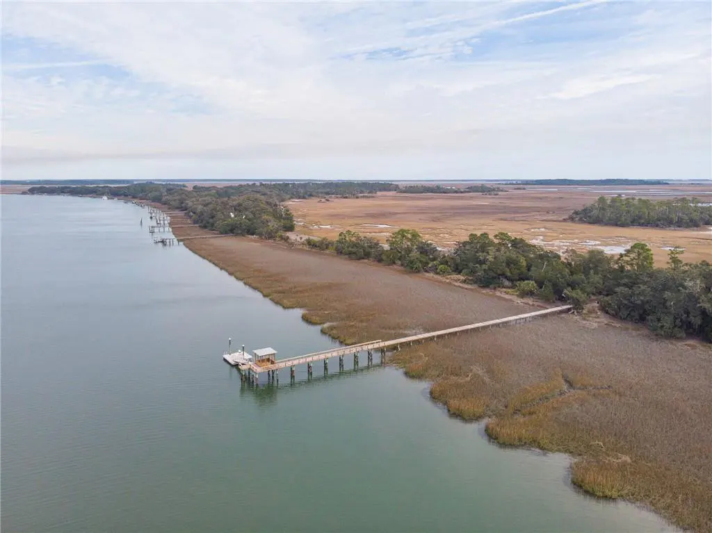Aerial view of a wooden dock extending into a river, surrounded by marshland and trees under a cloudy sky.