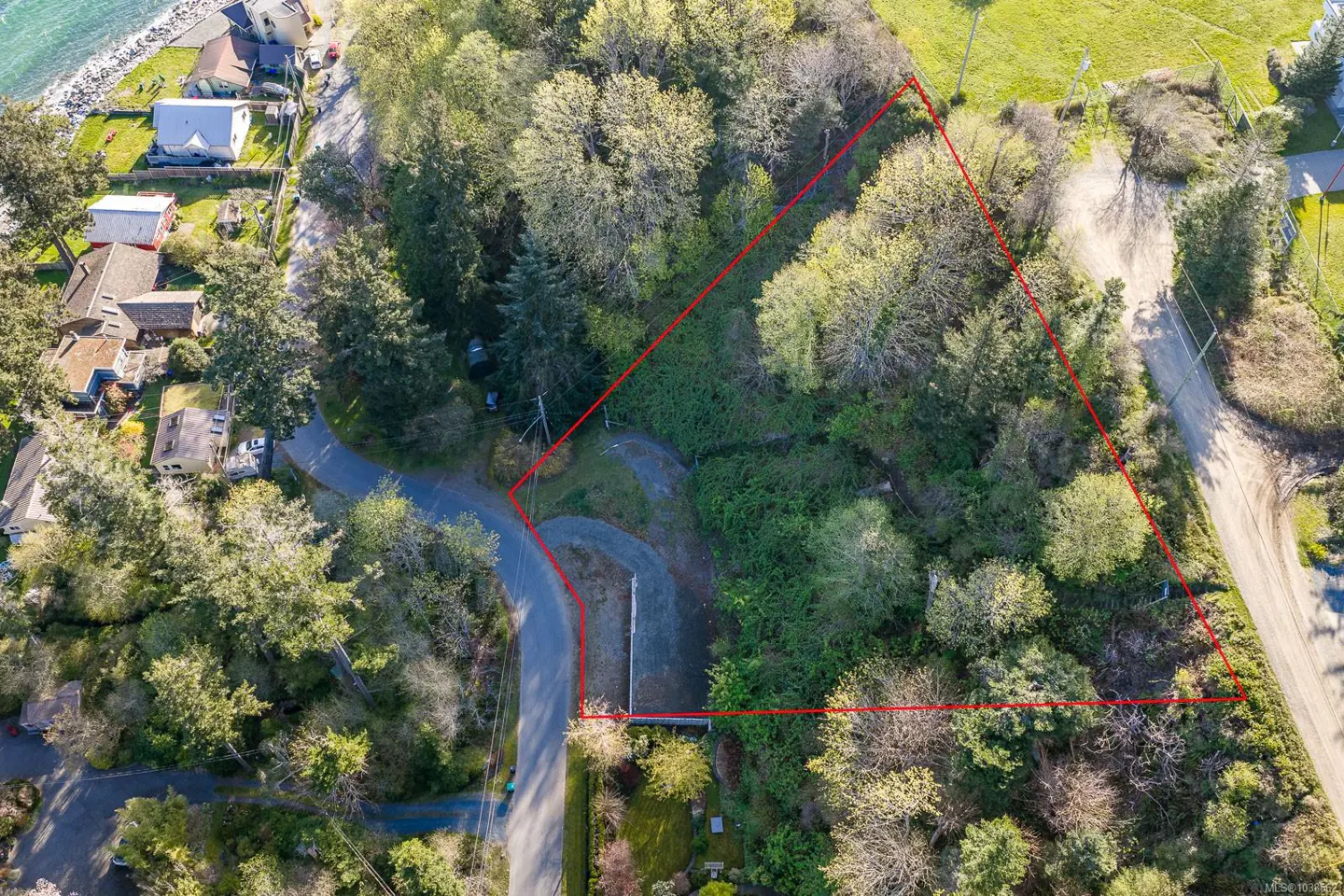 Aerial view of a wooded, triangular-shaped residential lot outlined in red, located near a coastal road and neighborhood.