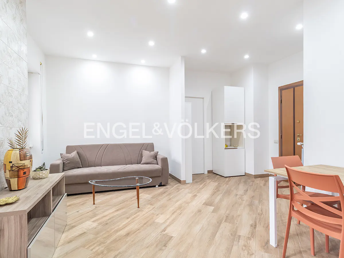Bright living room with a brown sofa, glass coffee table, and light wood floors. Orange chairs and a table are in the dining area.