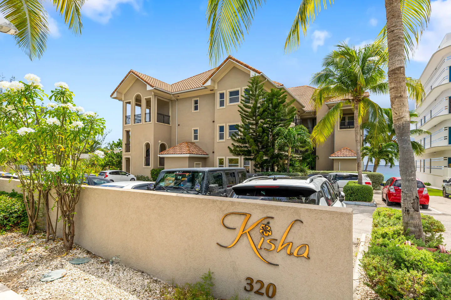 Exterior view of "Kisha 320" condos, a beige building with a red tile roof, palm trees, and parked cars.