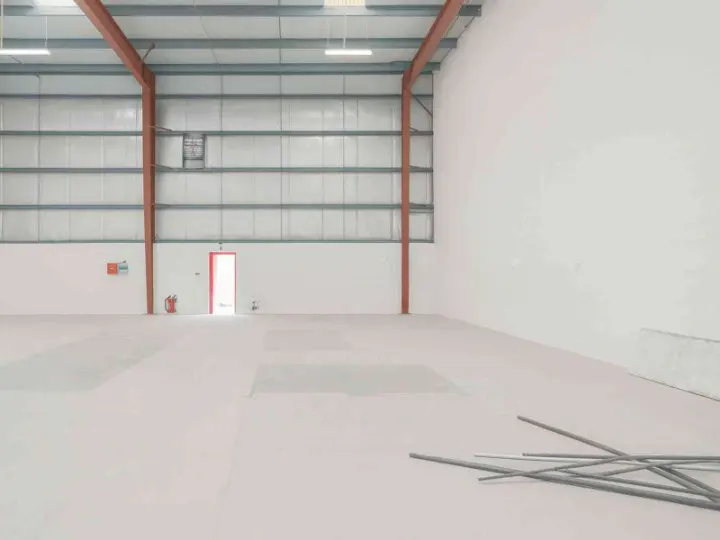 An empty warehouse with a white floor, white walls, and a metal ceiling. A red door and fire extinguisher are on the left wall.