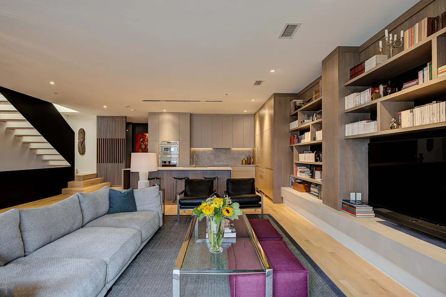 Interior view of a modern living room with a gray sofa, kitchen, and bookshelves. A glass coffee table with sunflowers sits on a gray rug.