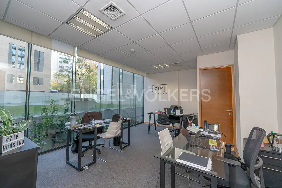 Office space with desks, chairs, and computers. Large windows offer a view of greenery and buildings. The Engel & Volkers logo is visible on the wall.