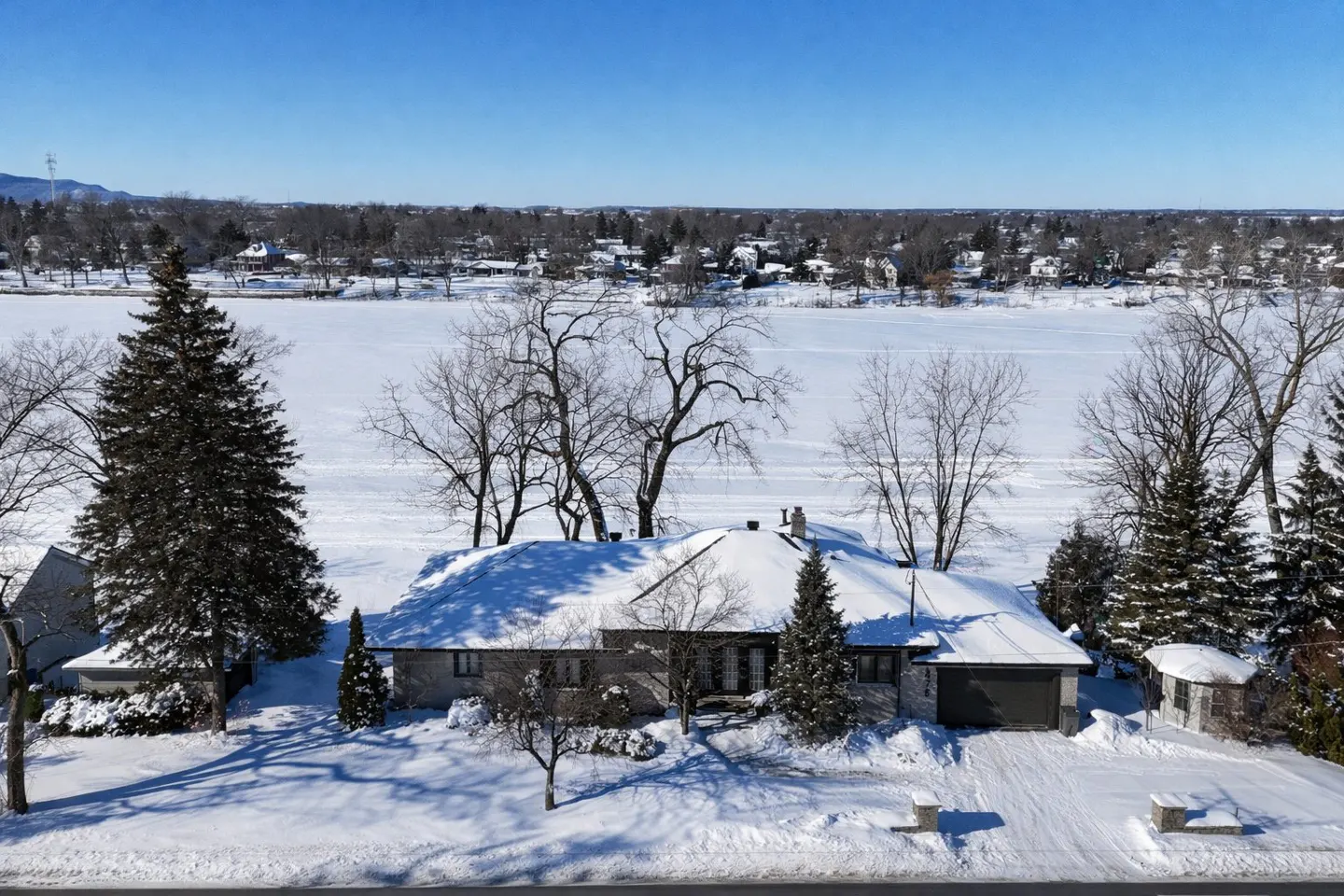 Aerial view of a snow-covered single-story house with a frozen lake and trees in the background on a sunny day.