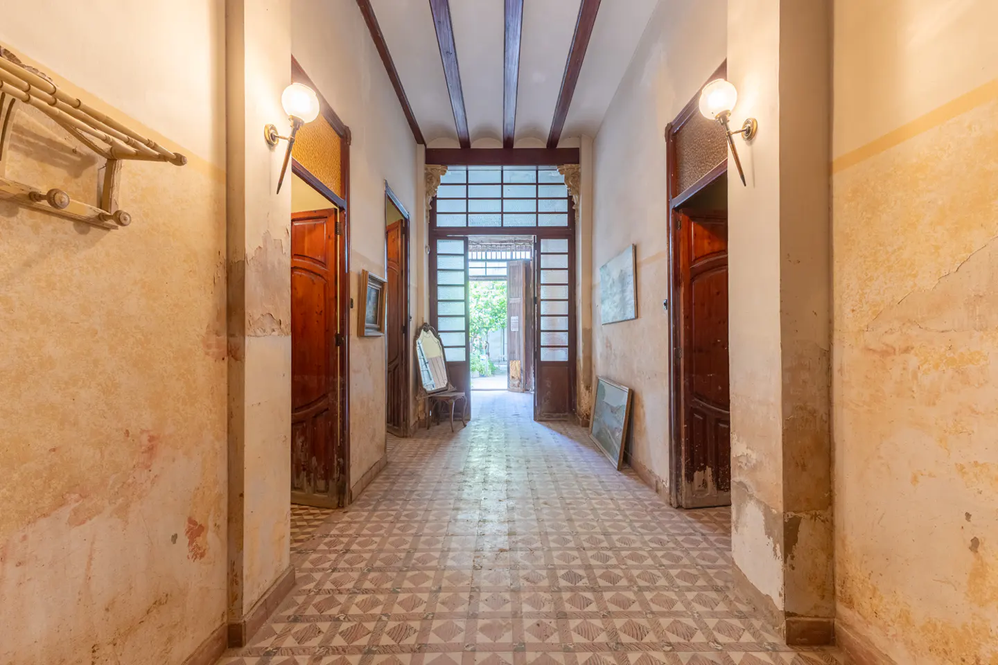 A rustic hallway with patterned tile floors, weathered beige walls, dark wood doors, and a glass-paneled door to a garden.