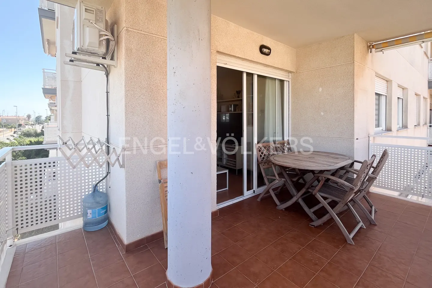 Balcony with a round wooden table and chairs, sliding glass doors, and a white railing. Brown tile floor. Engel & Völkers logo.