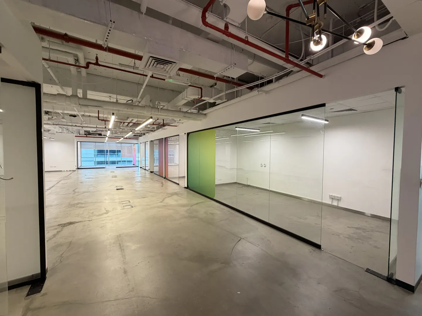 Empty office space with glass-walled rooms, concrete floors, and exposed ceiling pipes. A modern chandelier hangs in the foreground.