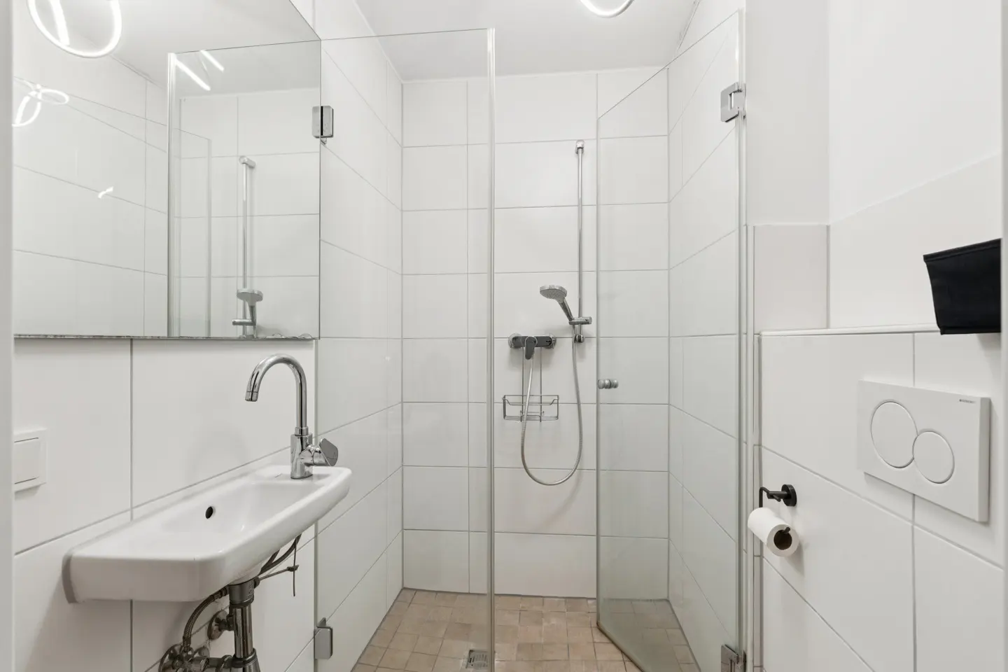 Bright bathroom with white tiled walls, a glass-enclosed shower, and a white sink with a chrome faucet. A toilet paper holder is mounted on the wall.