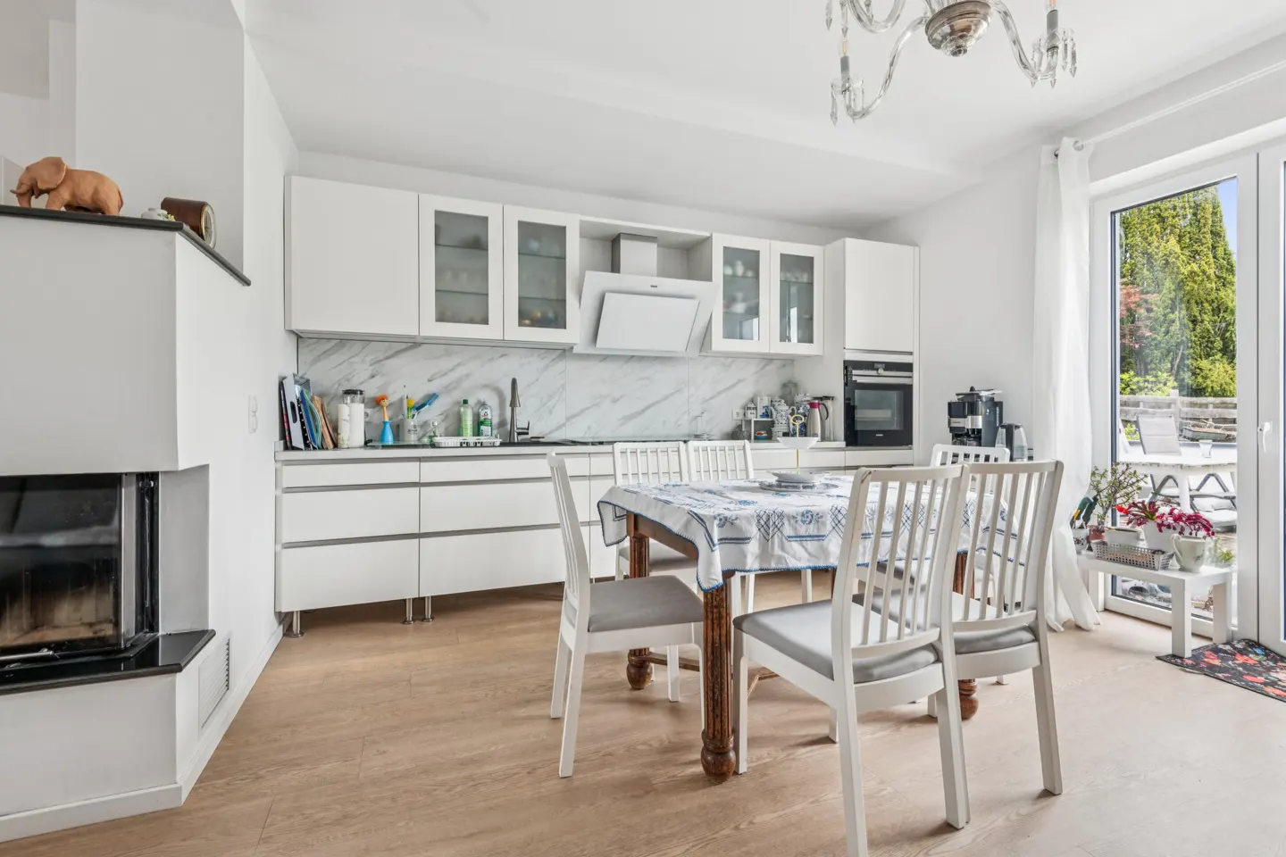 Bright kitchen with white cabinets, marble backsplash, and wood floors. A dining table with white chairs sits in the center. A fireplace is on the left.