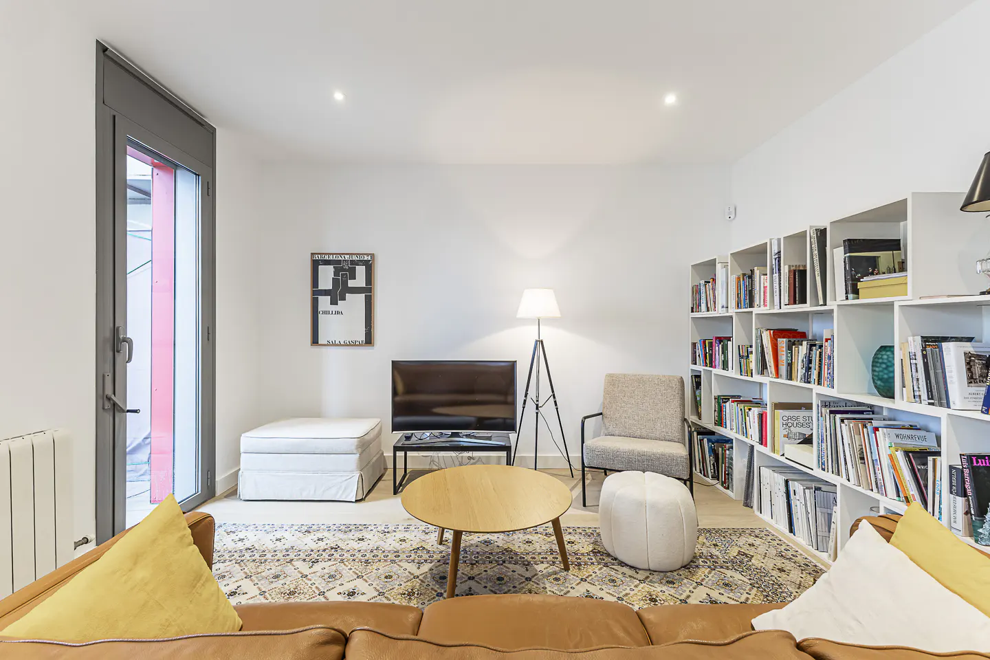 Bright living room with a brown leather couch, yellow pillows, a patterned rug, and a white bookshelf filled with books.
