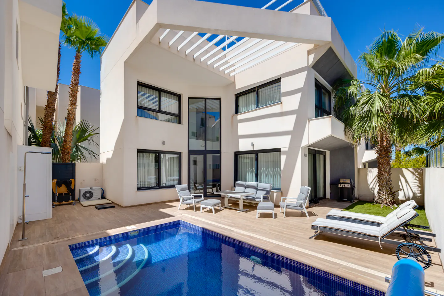Modern white house with a blue tiled pool, outdoor seating, and palm trees under a bright blue sky.