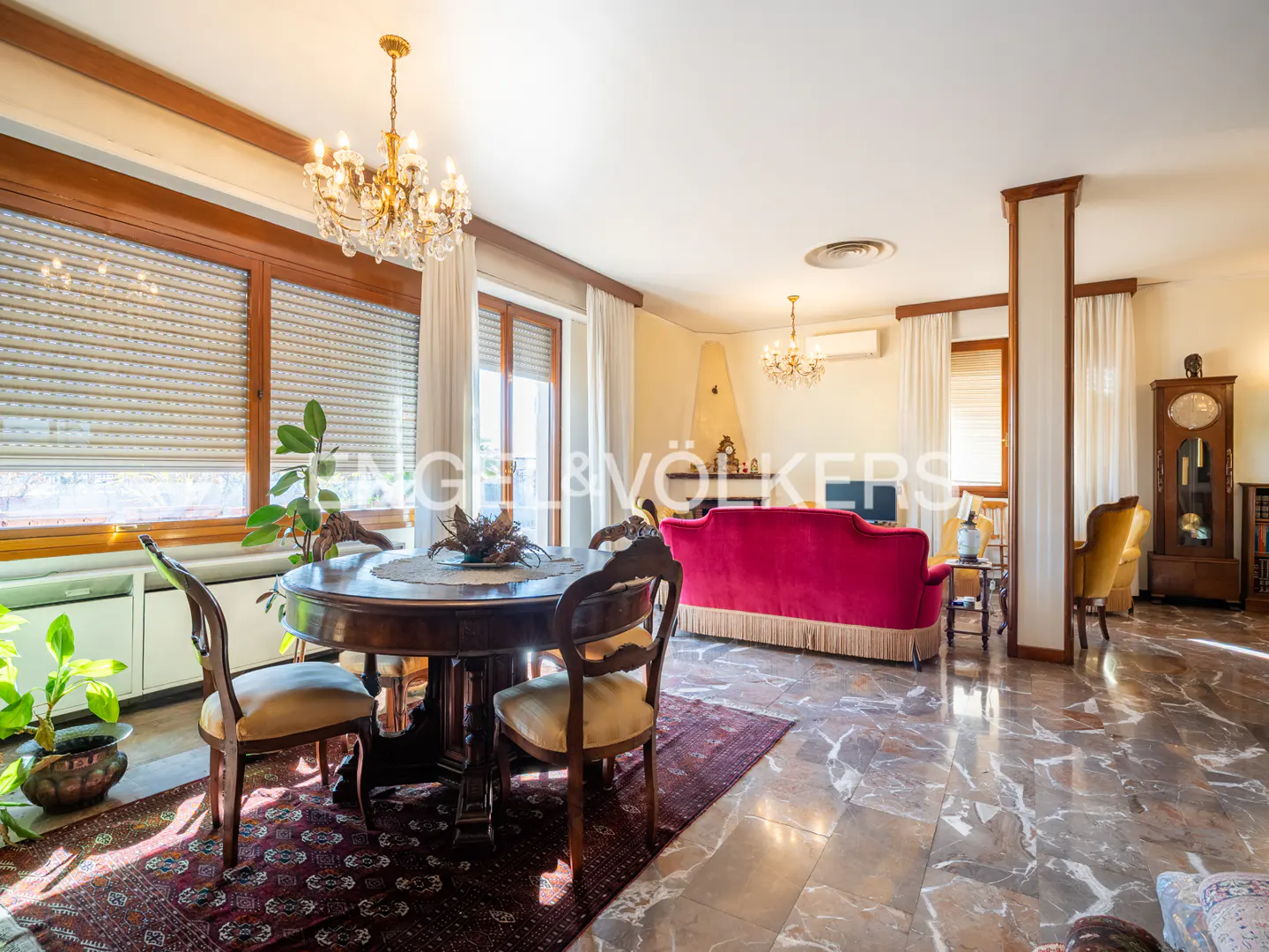 A bright, open-concept living and dining area with marble floors, a red sofa, and a round wooden table with chairs. Chandeliers hang from the ceiling.