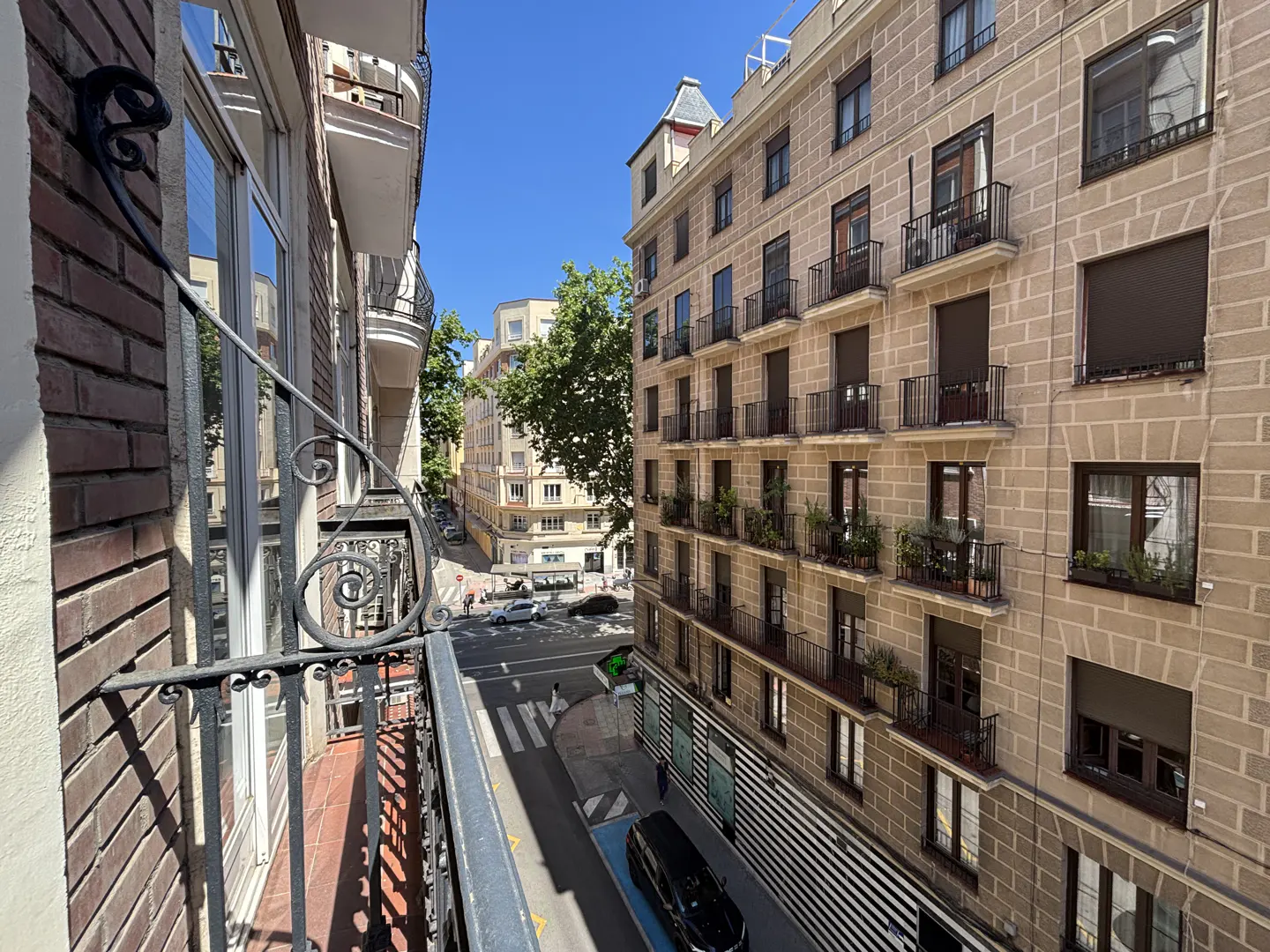 View from a balcony with ornate iron railings overlooking a city street with buildings and a crosswalk on a sunny day.