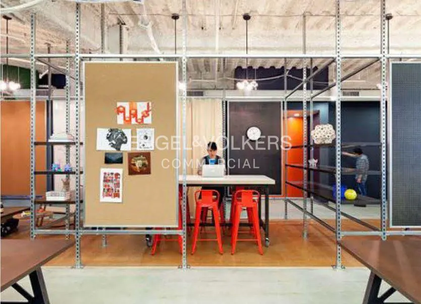 Office space with a woman working at a table with red chairs. A bulletin board with artwork is on the left. Shelving units divide the space.