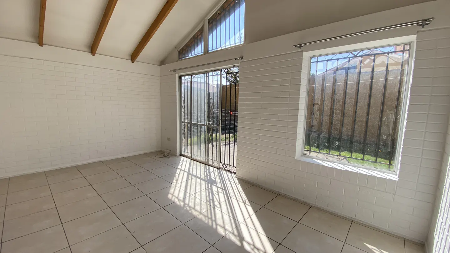 Bright, empty room with white brick walls, tile floor, and wooden ceiling beams. A barred window and door let in sunlight.